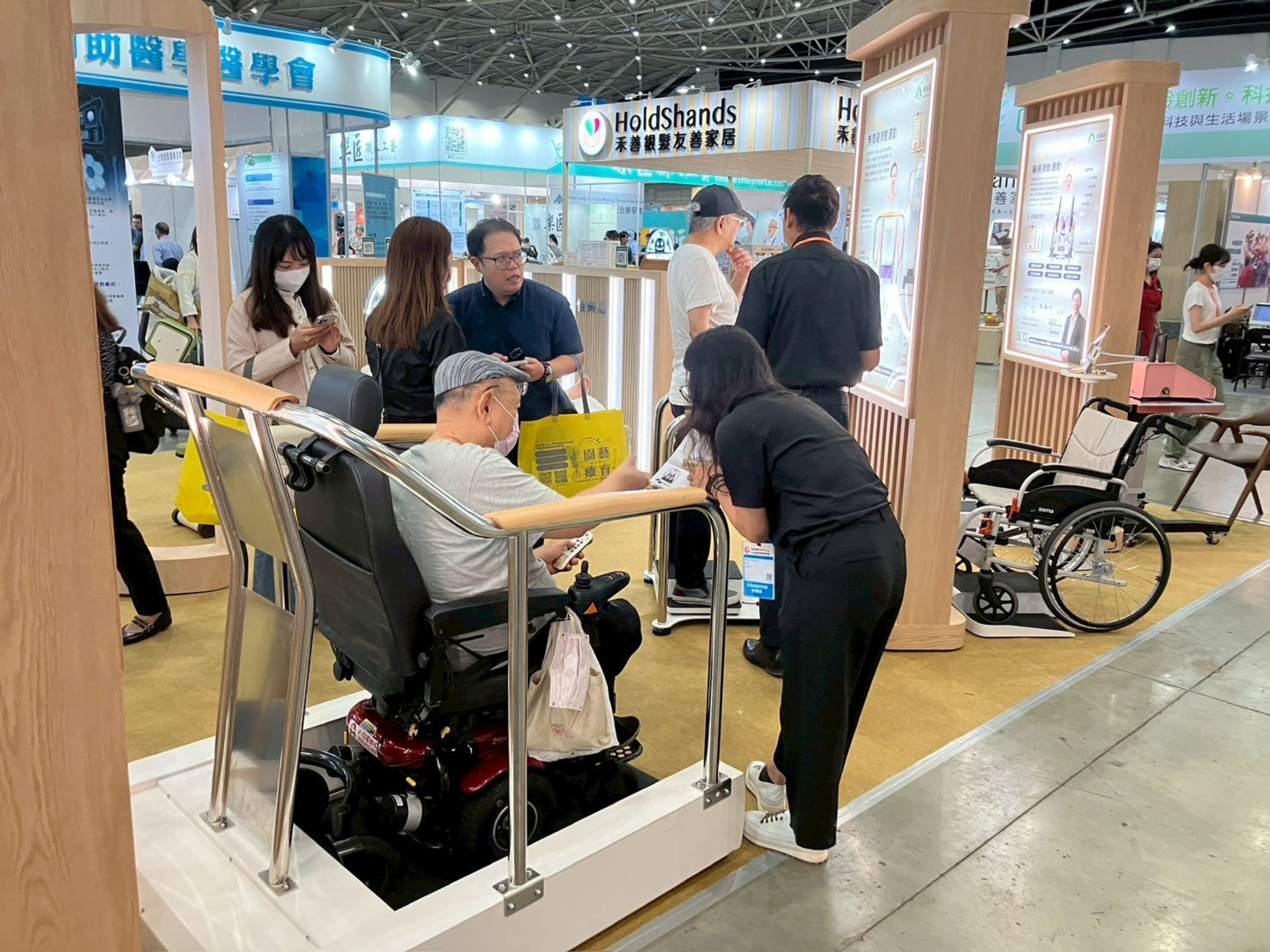 第六屆台北國際照顧博覽會特多勁展位。一位坐在電動輪椅上的長者正在體驗全球首創的無障礙律動平台/輪椅運動設備。特多勁專業團隊正彎腰進行操作指導和解說，展現無障礙運動解決方案。